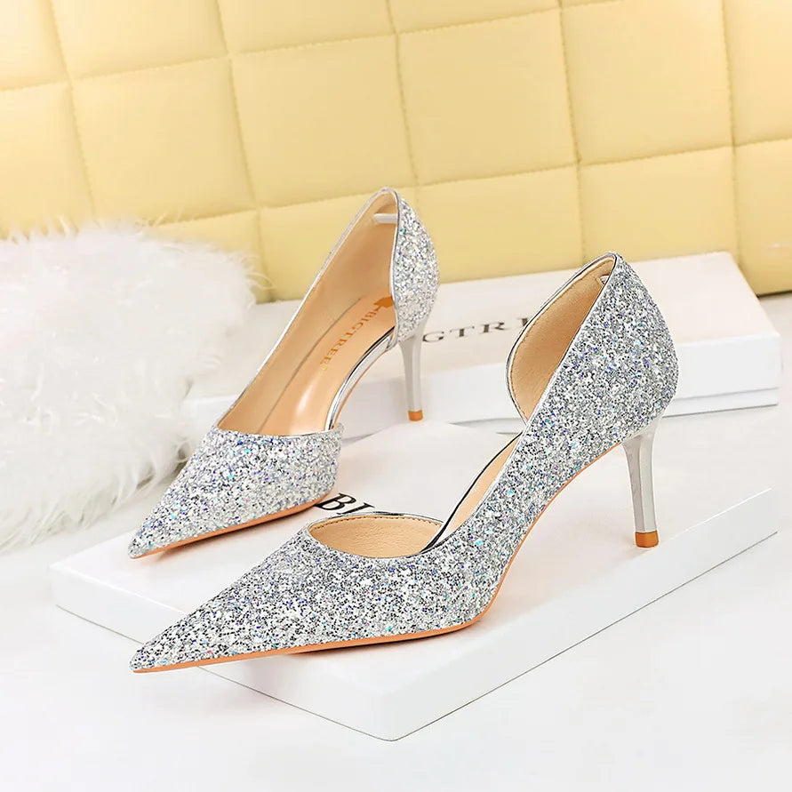 Silver glitter high heels with pointed toes and mid-height stiletto heels, placed on white boxes against a quilted yellow backdrop and fluffy white rug.