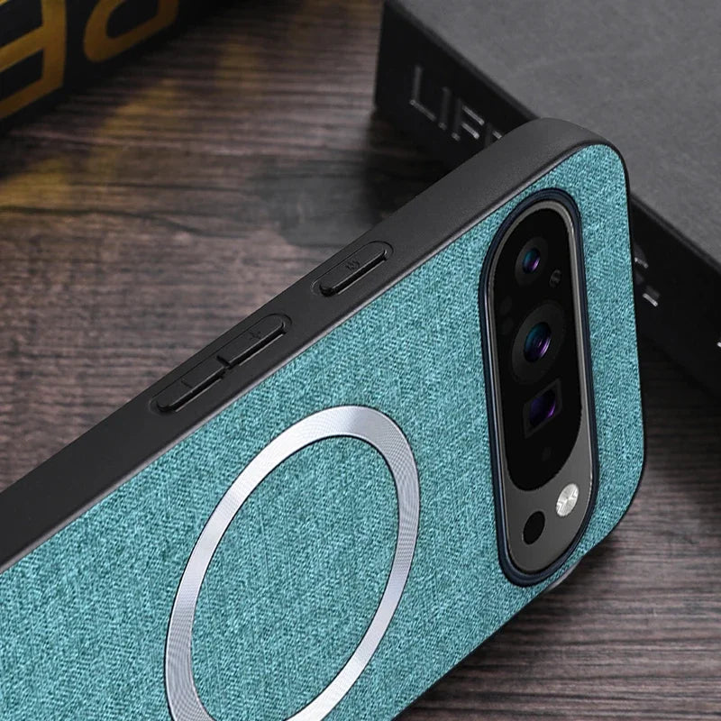 Close-up of a smartphone with a teal fabric case featuring a metallic ring and dual camera setup. The phone is placed on a dark wooden surface with blurred background objects.