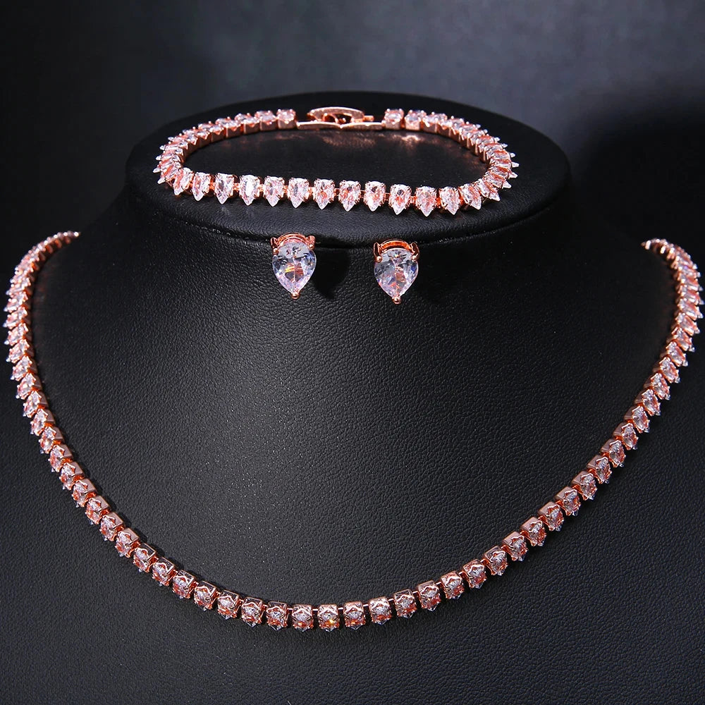 Rose gold jewelry set with clear stones includes a sparkling necklace, a matching bracelet, and teardrop-shaped earrings, displayed on a black stand.