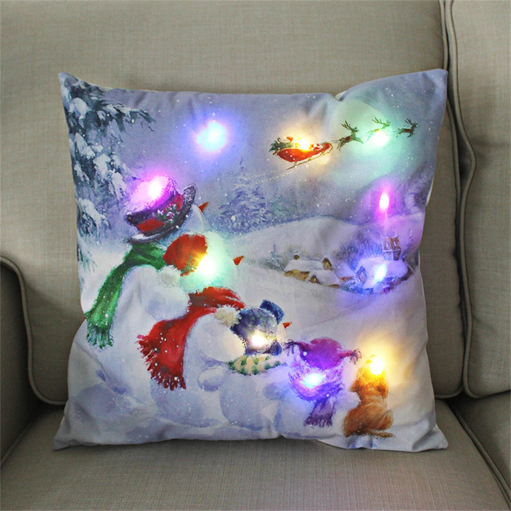 Festive pillow with snowmen, scarves, and a snowy village scene. Features colorful LED lights, including a glowing Santa's sleigh, enhancing the winter holiday theme.