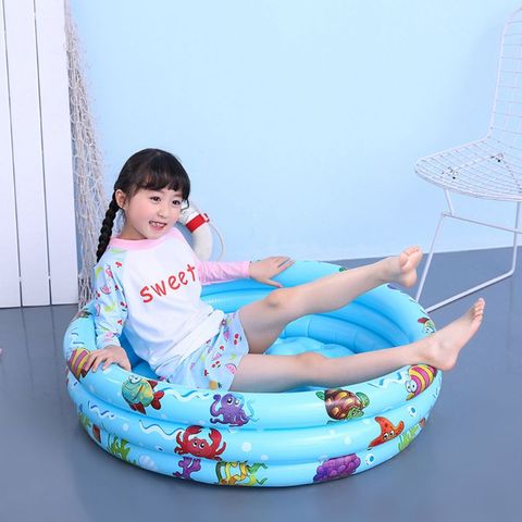 A young girl wearing a white shirt with "sweet" written on it and colorful shorts sits playfully in a small, blue inflatable pool decorated with cartoon sea creatures, against a light blue wall.