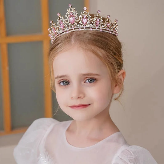 Young girl with blonde hair wears a pink jeweled tiara and a white dress, smiling softly. She stands in front of a blurred background with a wooden window frame.