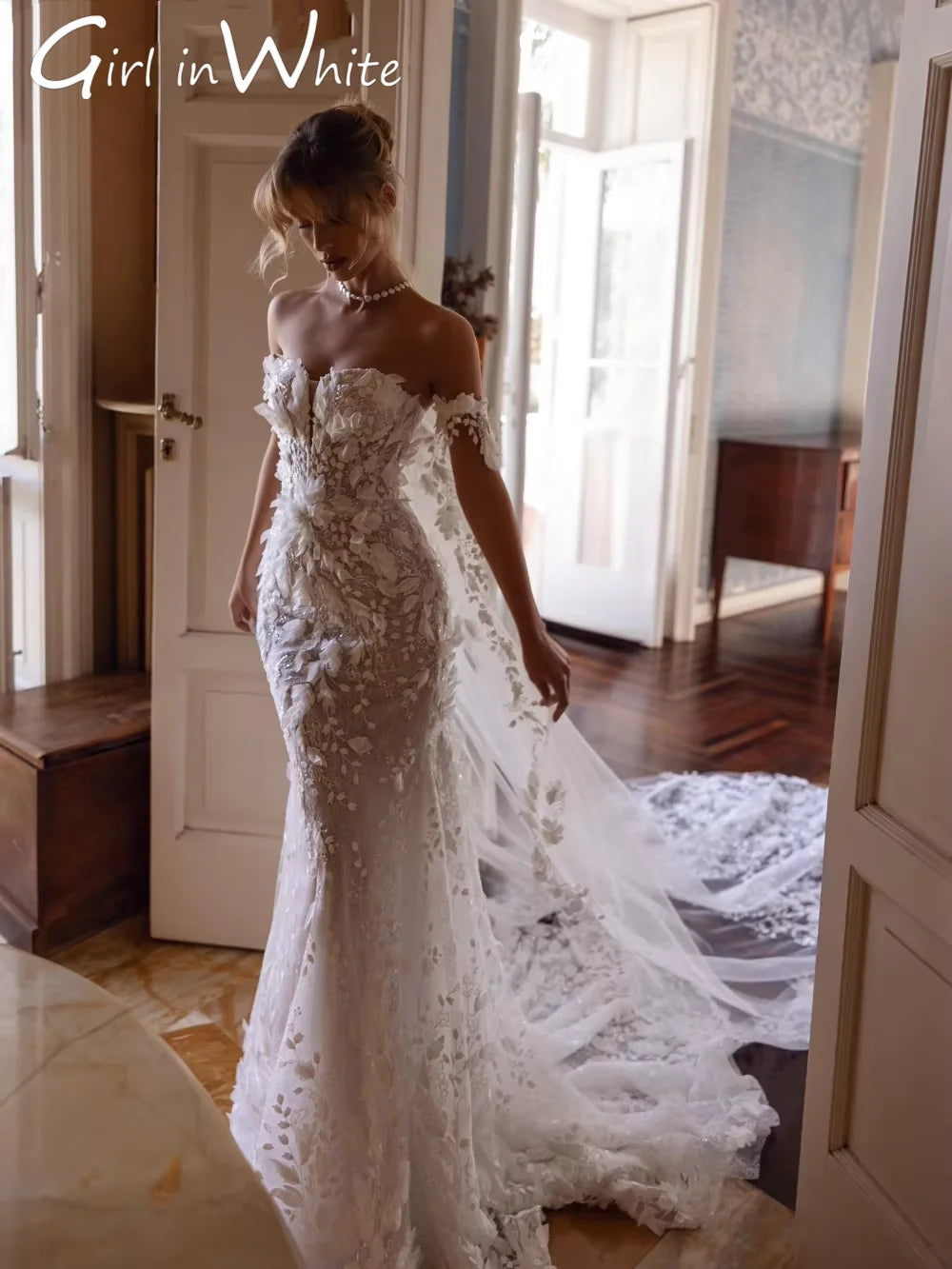 A woman in an ornate, off-shoulder, lace wedding dress with a long train, stands gracefully near an open door in a softly lit room with wooden floors and patterned walls.