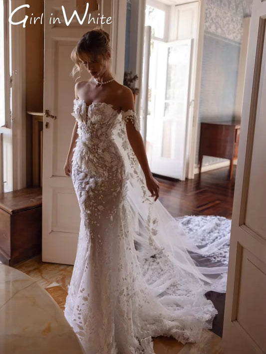 A woman in an ornate, off-shoulder, lace wedding dress with a long train, stands gracefully near an open door in a softly lit room with wooden floors and patterned walls.