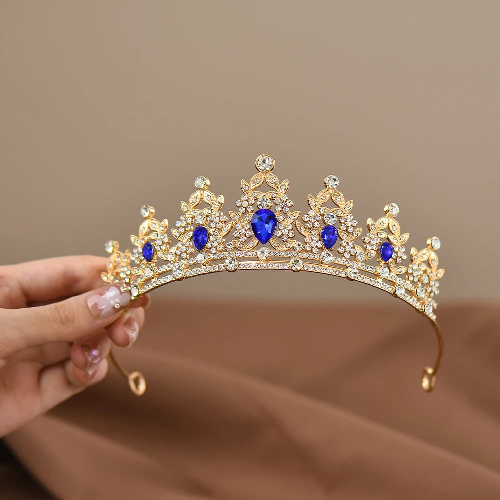 A gold tiara adorned with blue teardrop gemstones and clear rhinestones is held by a hand with manicured nails against a blurred brown background.