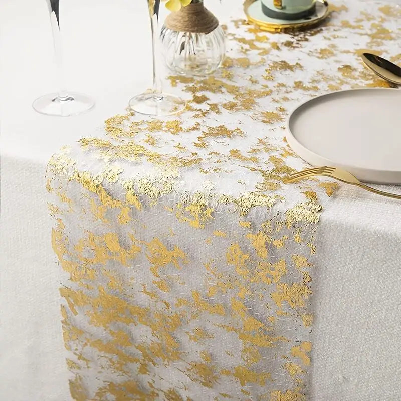 Elegant table setting with a white and gold abstract table runner. Includes a gray plate, gold cutlery, glasses, and a small decorative vase on a white tablecloth.