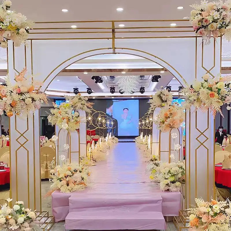Elegant wedding aisle adorned with pastel floral arrangements and golden arches, leading to a stage with a large screen displaying an image, in a lavishly decorated venue.