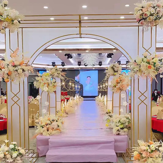 Elegant wedding aisle adorned with pastel floral arrangements and golden arches, leading to a stage with a large screen displaying an image, in a lavishly decorated venue.