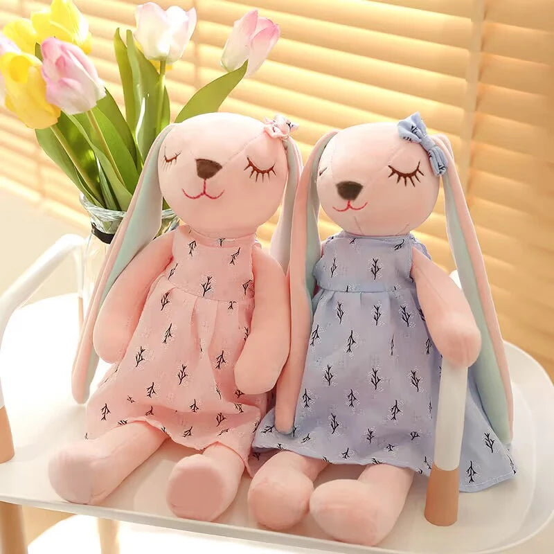 Two plush bunny toys with closed eyes wear pink and blue dresses with a delicate pattern, sitting on a white surface. A vase of tulips is in the background.