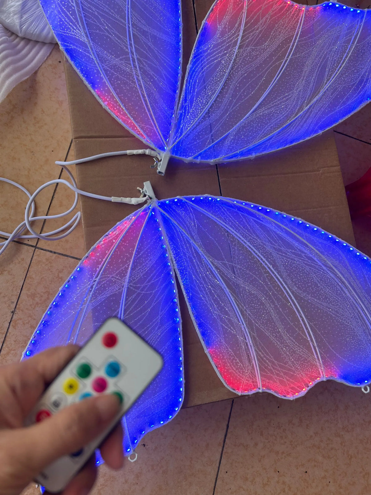 LED-illuminated butterfly wings with blue and red lights on a cardboard surface, controlled by a remote with colorful buttons held in a hand.