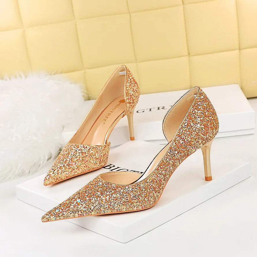 Gold glitter high-heeled shoes with pointed toes on white boxes, set against a soft cream quilted background and a fluffy white rug accent.