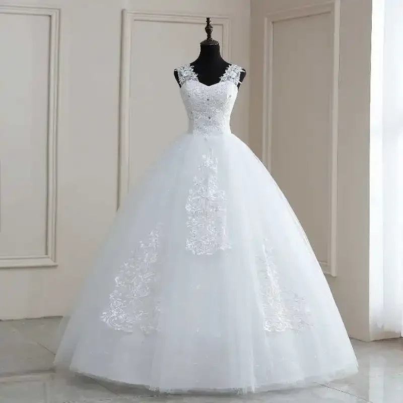 A white ball gown wedding dress on a mannequin, featuring an embroidered lace bodice and straps. The full skirt has floral lace appliques, set in a bright, elegant room.