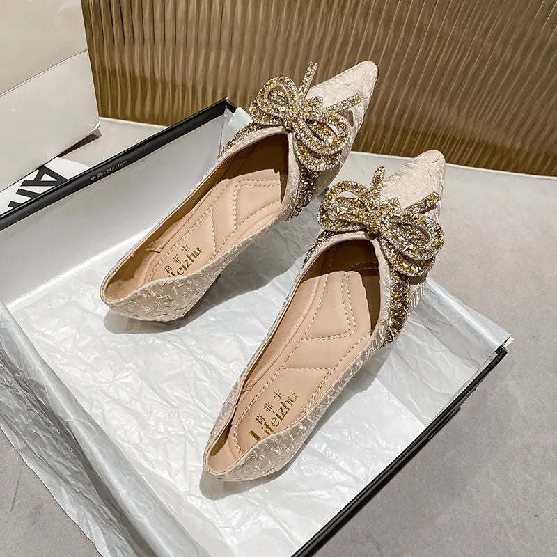 Elegant cream-colored pointed flats with ornate gold sequin bows on top, displayed in an open shoe box lined with white tissue paper on a light-colored surface.