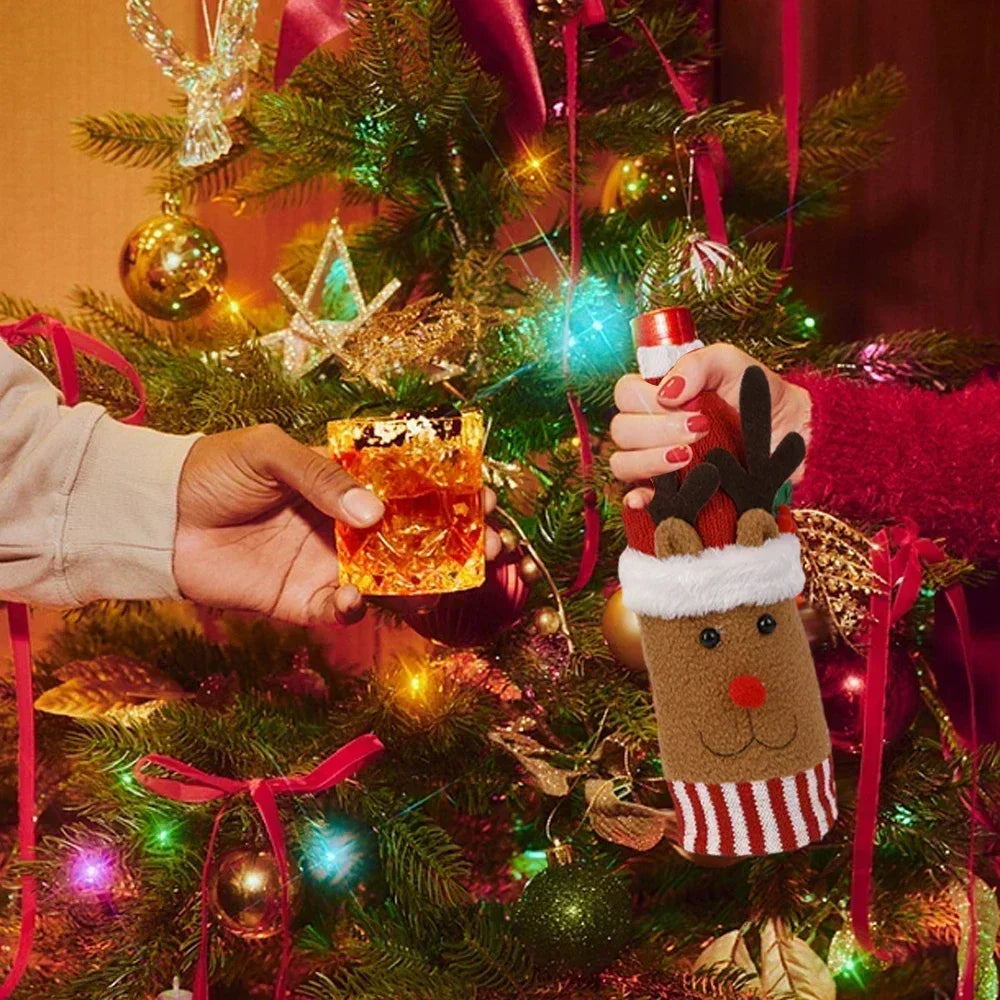 Hands exchanging a festive drink by a decorated Christmas tree. One holds a cocktail, the other a bottle in a reindeer cozy. The tree is adorned with lights and ornaments.