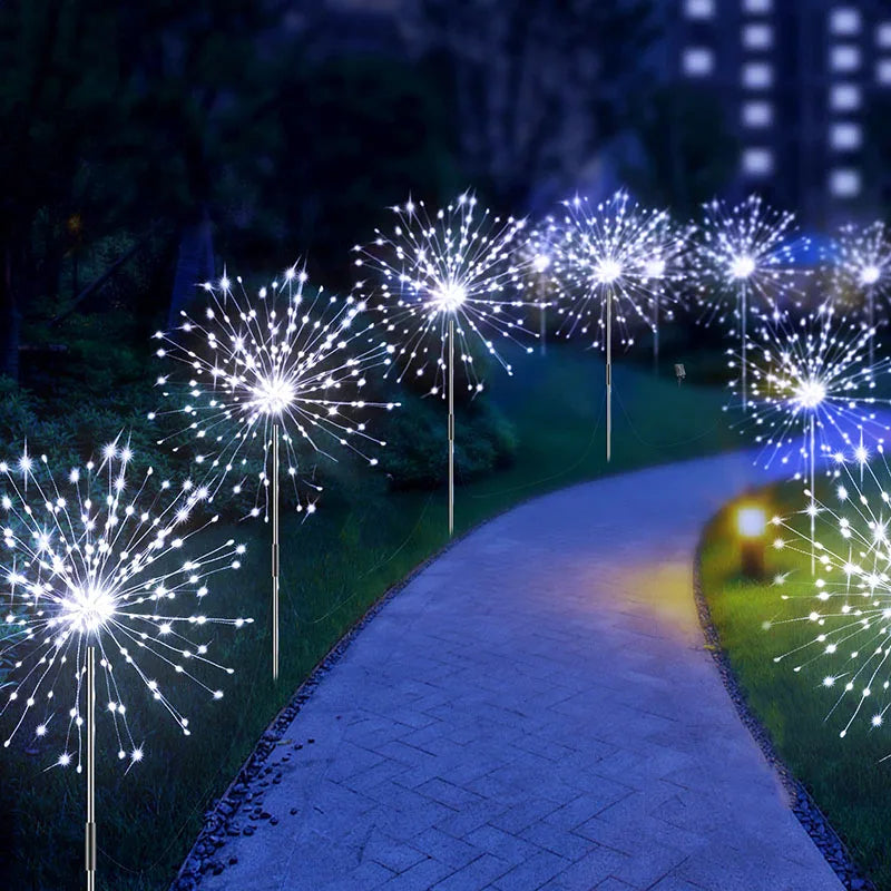 A nighttime garden scene with a winding path illuminated by dandelion-shaped LED lights that resemble fireworks, creating a whimsical and enchanting atmosphere.