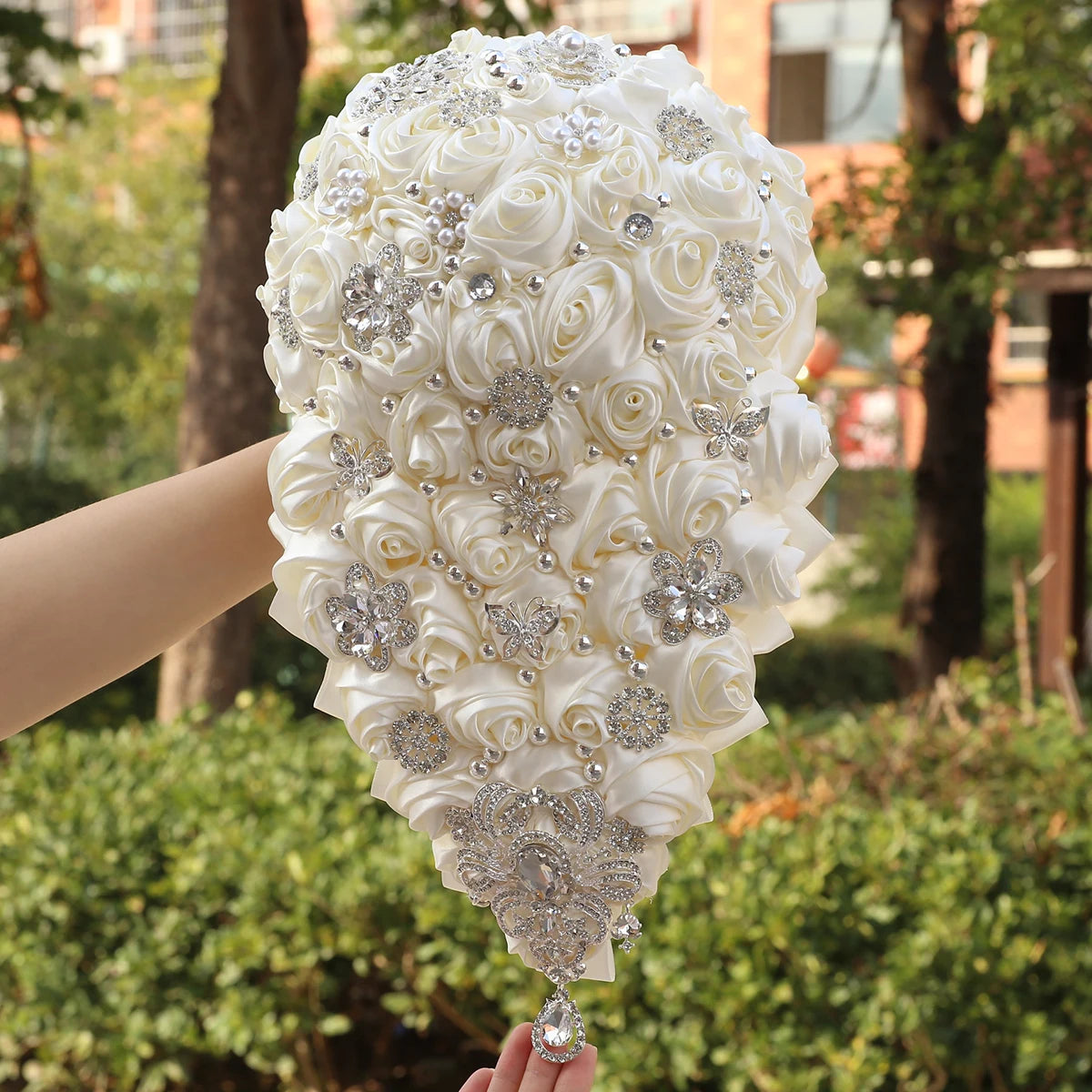A teardrop bridal bouquet with white satin roses, adorned with sparkling rhinestones, pearls, and intricate silver embellishments, held against a garden background.