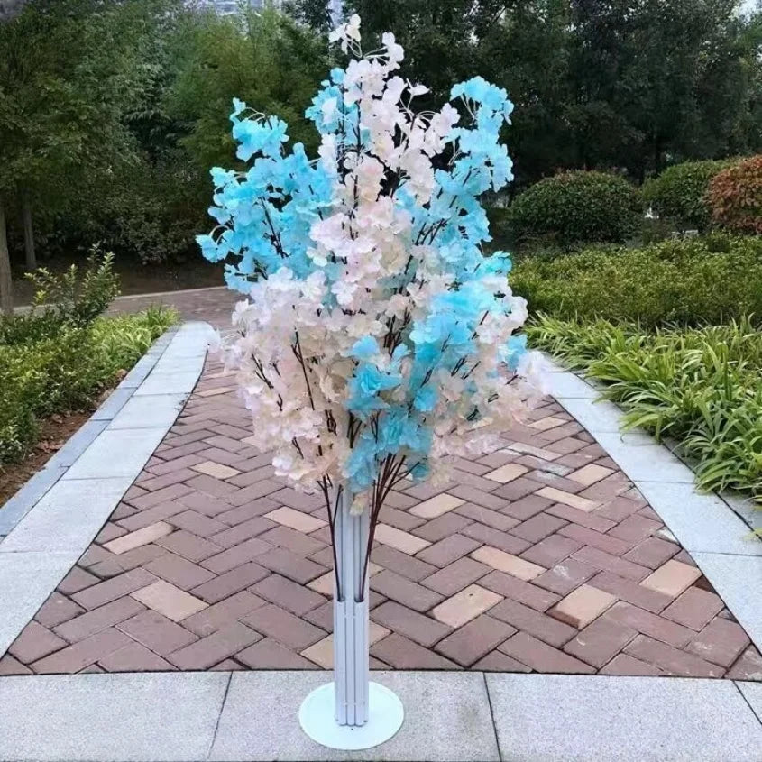 A decorative arrangement of blue and white artificial flowers on a stand is placed on a paved walkway, surrounded by lush greenery in a garden setting.