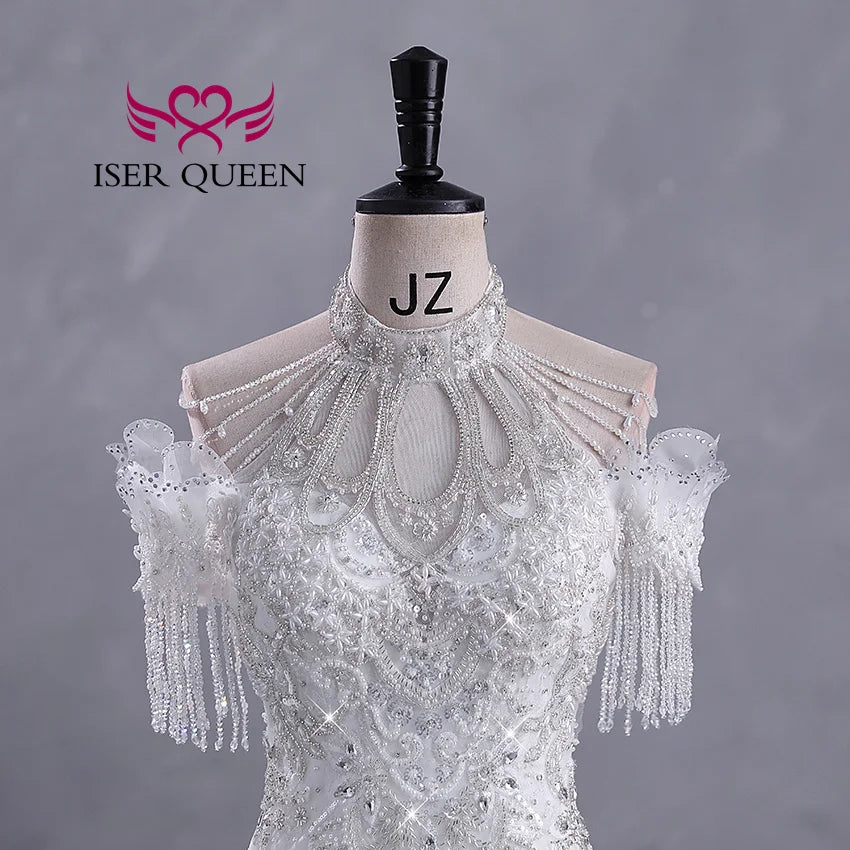 Elegant white wedding dress on mannequin with intricate beadwork, lace detailing, and pearl accents. Shoulder design features draped pearls and sequined fringe. Background is gray.