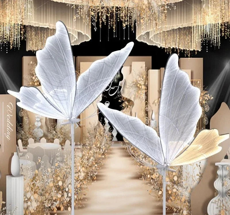 Two large, translucent butterfly decorations with delicate wings stand in a luxurious wedding setting, adorned with gold and floral elements, under a chandelier canopy.