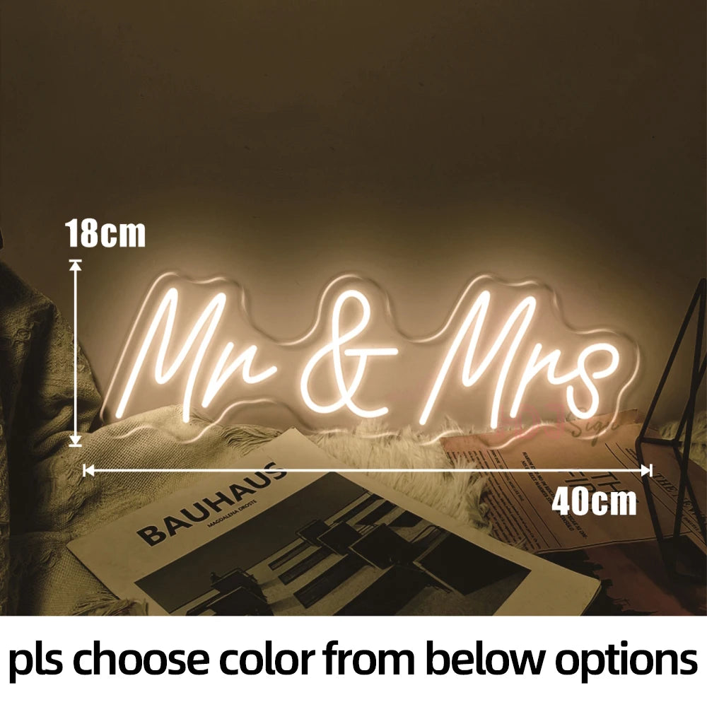 Neon sign reading "Mr & Mrs," measuring 18cm by 40cm, emits a warm glow. It's placed on a textured surface beside magazines, with a choice of colors available.