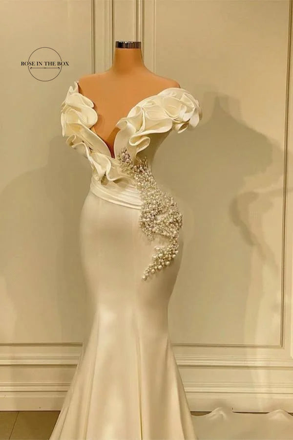 Elegant white gown on display with sculpted shoulders, floral embellishments, and pearl accents. The silhouette is fitted, flaring at the bottom, displayed against a neutral backdrop.