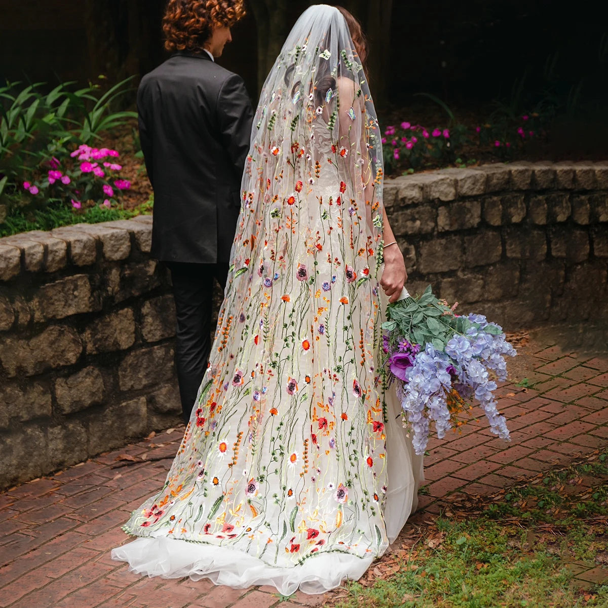 A bride in a floral embroidered gown and veil holds purple flowers. She stands on a brick path next to a groom in a black suit, near a stone wall and vibrant garden.