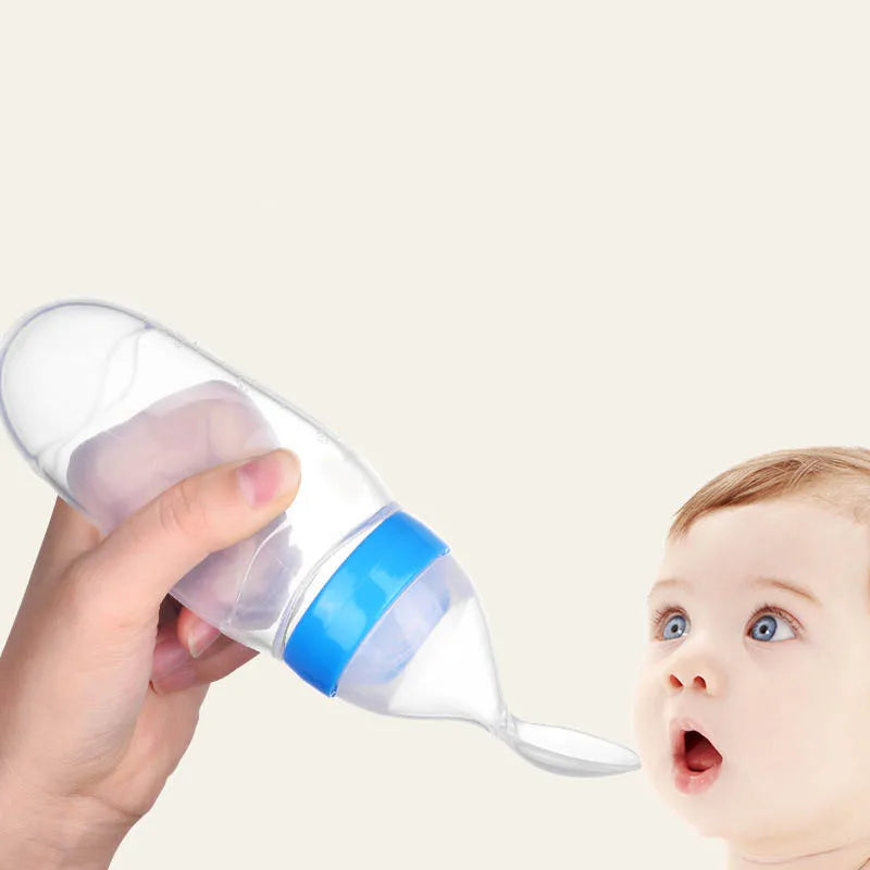 A hand holds a baby feeding bottle with a spoon tip, approaching an infant with wide eyes and an open mouth against a plain background.