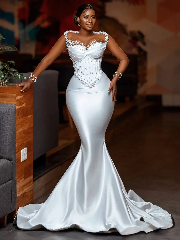 Woman in an elegant, form-fitting white gown with a pearl-embellished corset top, standing confidently in a stylish interior with warm lighting and modern decor.