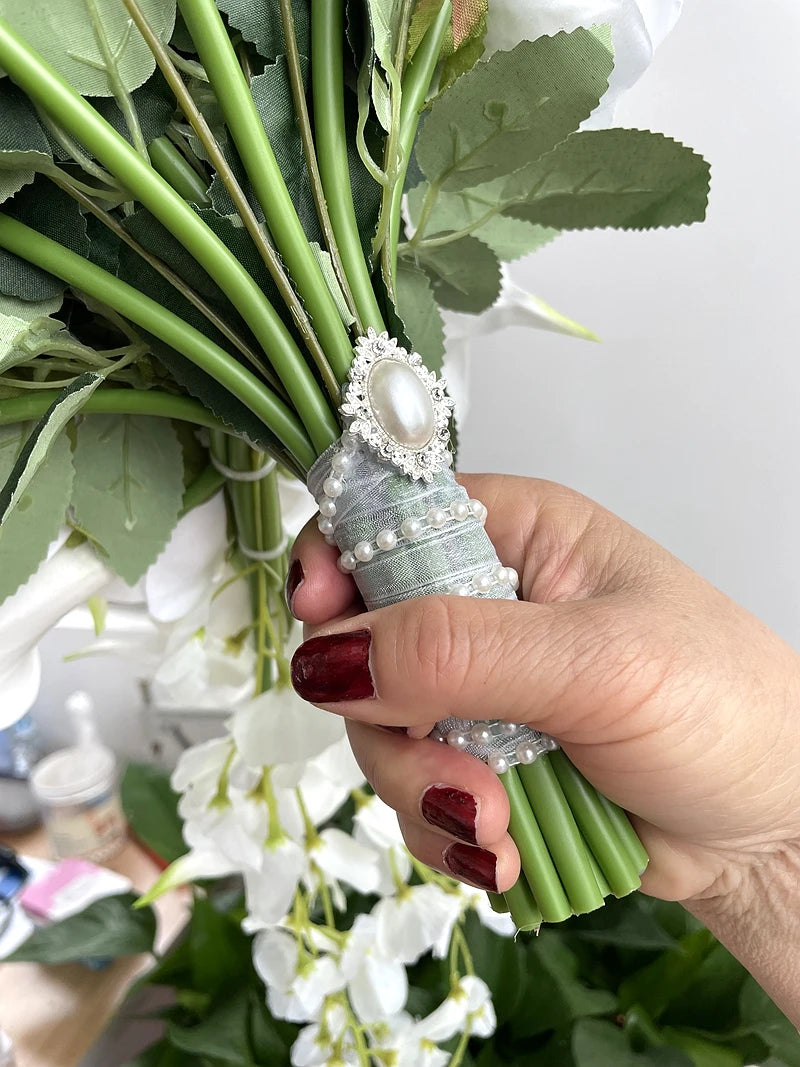 A hand with dark red nails holds a bouquet of green stems wrapped in silk adorned with pearl beads and a decorative pearl brooch. White flowers and green leaves are visible.