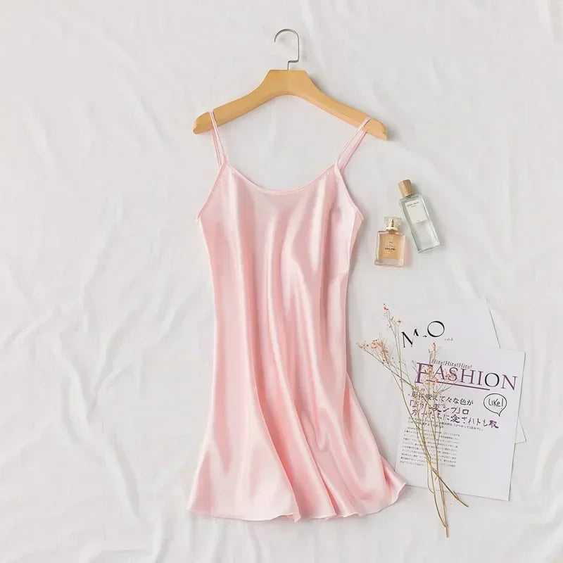 A silky pink slip dress on a hanger rests on a white background, accompanied by two perfume bottles, fashion magazines, and a small sprig of delicate flowers.