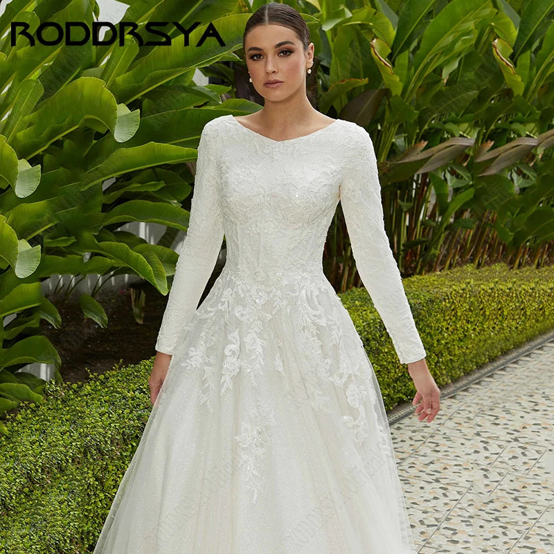 A woman in a long-sleeve, lace-embroidered white wedding dress stands on a paved path, surrounded by lush green foliage, displaying elegance and grace.