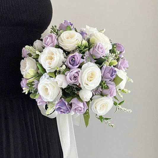 A person holds a bouquet of white and lavender roses with greenery, tied with a white ribbon, against a plain light background, contrasting with their dark clothing.