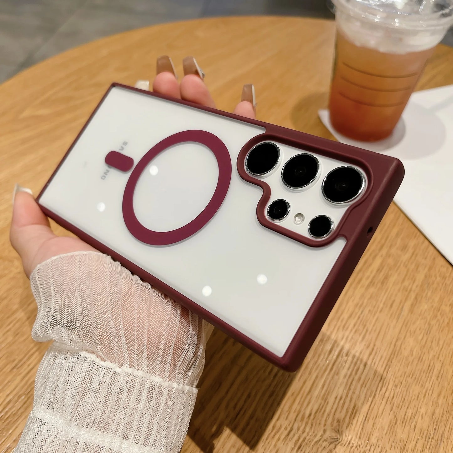 Hand holding a smartphone with a burgundy and white case featuring a circular design and multiple camera lenses. Background shows a wooden table with a cold drink.