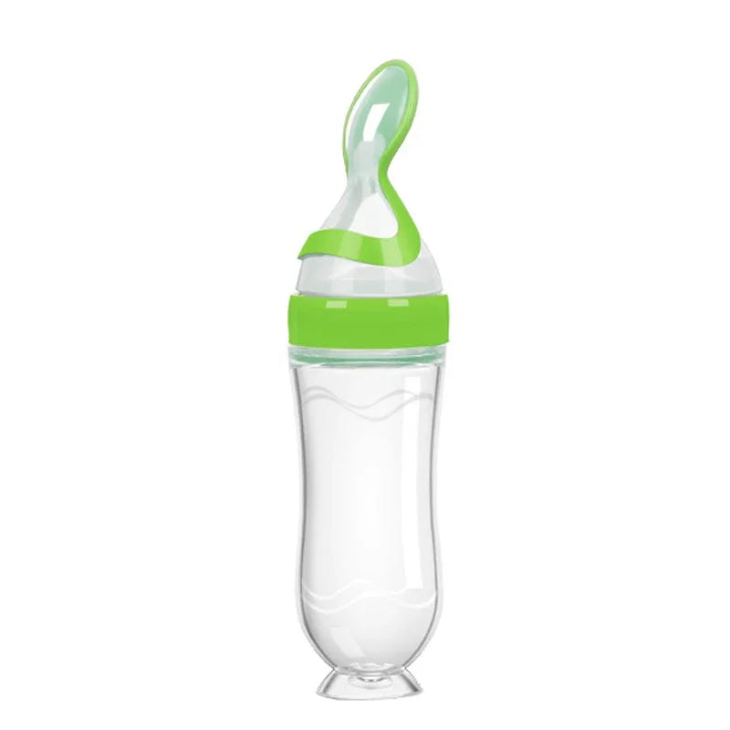 Transparent baby bottle with a green lid, designed with an integrated silicone spoon for easy feeding. The bottle has a curved shape and a wide base for stability.