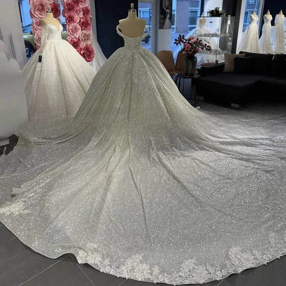 Luxurious bridal showroom with two elegant, sparkling wedding gowns on display. Each dress features intricate detailing, one adorned with lace trim, set against a backdrop of large pink roses.