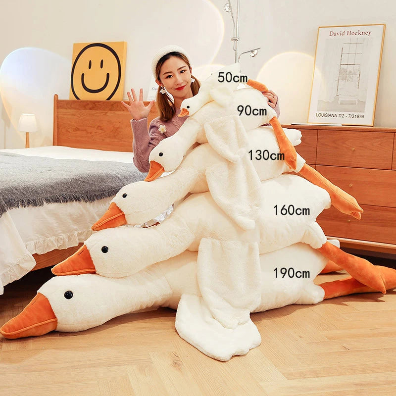 A woman sits next to a stack of five plush geese of varying sizes labeled 50cm to 190cm in a cozy bedroom, smiling and waving at the camera.