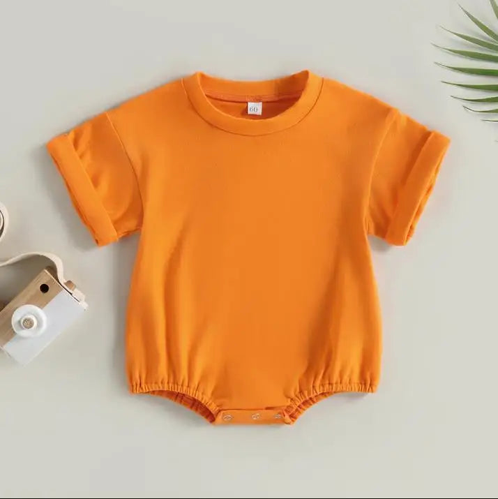 Orange baby bodysuit with short sleeves and snap buttons at the bottom, laid flat on a light background. A small wooden toy camera is on the left, with a sprig of leaves on the top right.