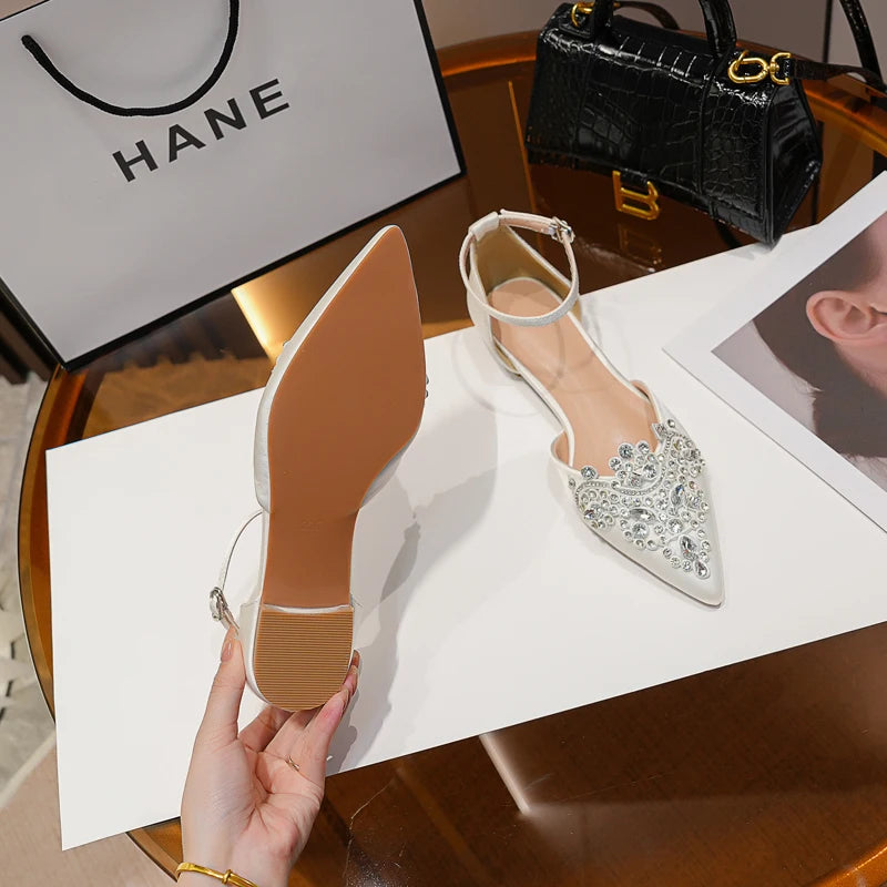 White pointed-toe flats adorned with rhinestones and ankle straps on a table, beside a black handbag and a shopping bag labeled "HANE." A hand holds one shoe, showing the sole.