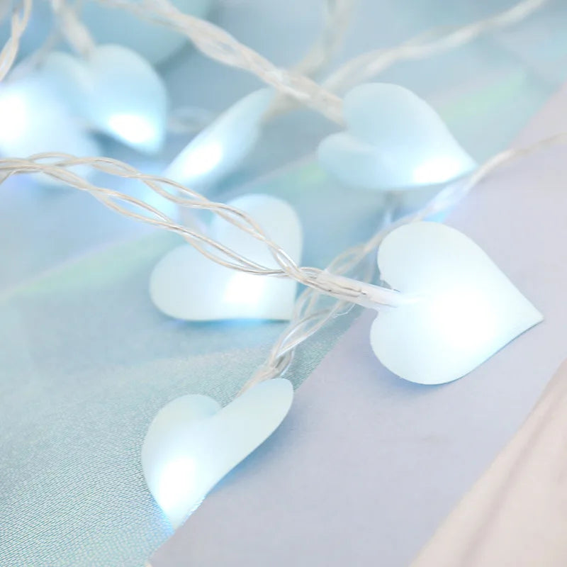 String lights with glowing white heart-shaped covers, intertwined on a soft, pastel blue and white surface. The scene evokes a gentle and romantic atmosphere.