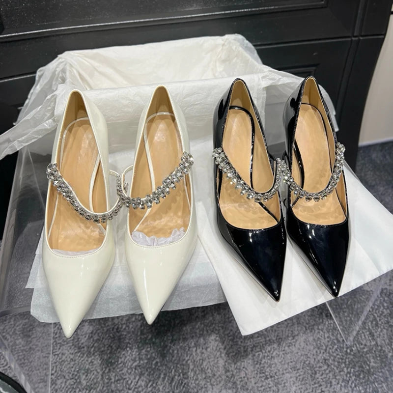 Two pairs of pointed-toe high heels with rhinestone ankle straps, one in glossy white and the other in glossy black, displayed on white tissue paper in a box.