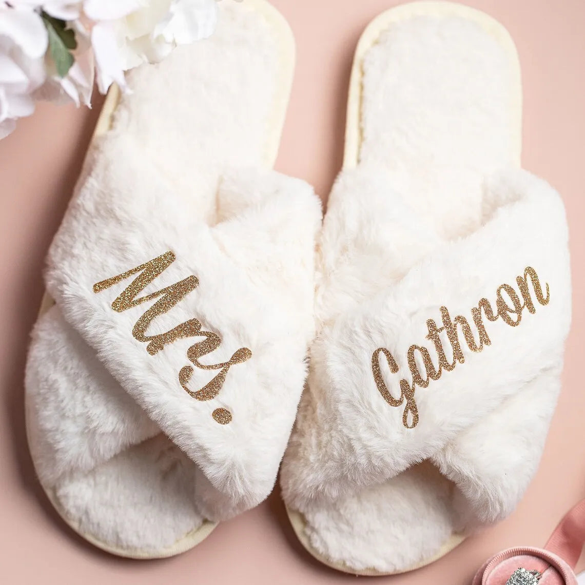 Fuzzy white slippers with gold glitter text reading "Mrs. Gathron." The slippers are on a soft pink surface, adorned with a pink ribbon and white flowers nearby.