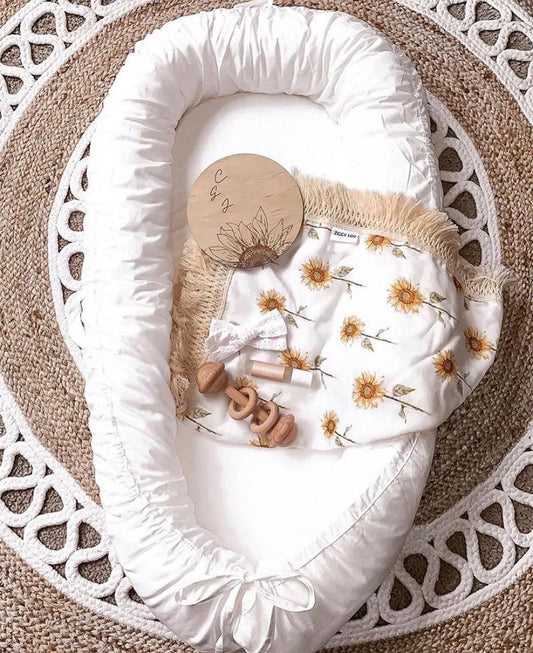 A cozy baby nest with a white ruffled edge, adorned with a sunflower blanket, wooden teether, and a round wooden plaque, set on a woven circular rug.