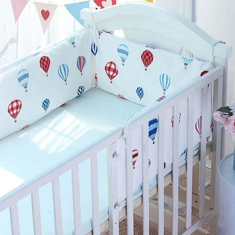 White crib with colorful hot air balloon print bedding. Light streams through a window decorated with heart and triangle flags, casting soft shadows. Pink flowers in a vase beside.