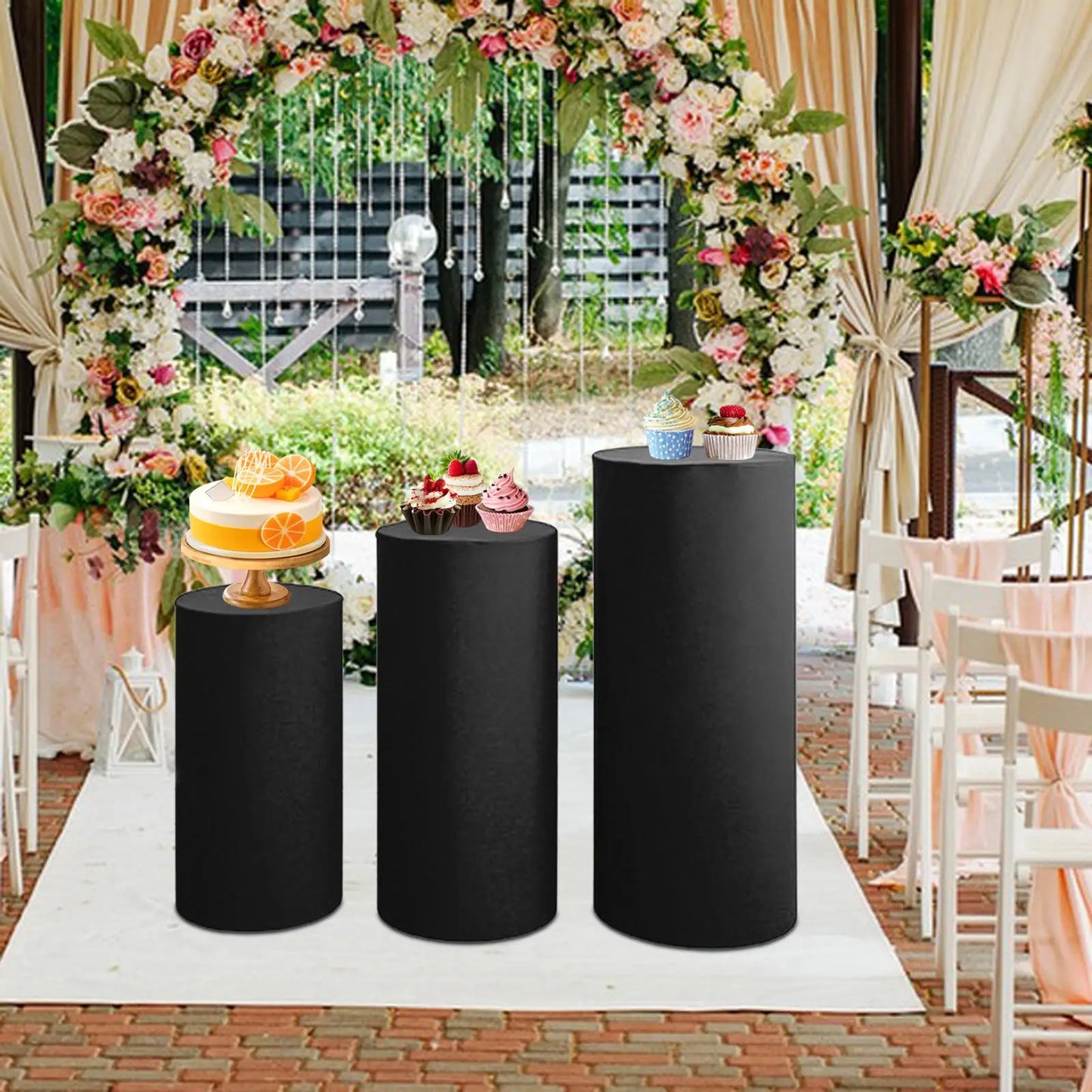 Dessert display with a cake and cupcakes on black stands, set in a floral-decorated outdoor wedding venue with white chairs and a brick pathway.