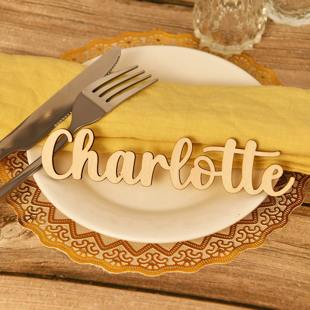 Place setting with a white plate on a wooden table, adorned with a yellow napkin, silverware, and a wooden name cutout "Charlotte". Brown doily adds a decorative touch.