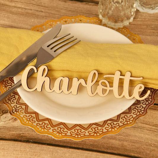 Place setting with a white plate on a wooden table, adorned with a yellow napkin, silverware, and a wooden name cutout "Charlotte". Brown doily adds a decorative touch.