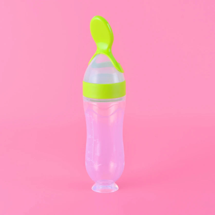 A clear baby bottle with a green spoon-shaped cap and silicone lid sits upright against a pink background.