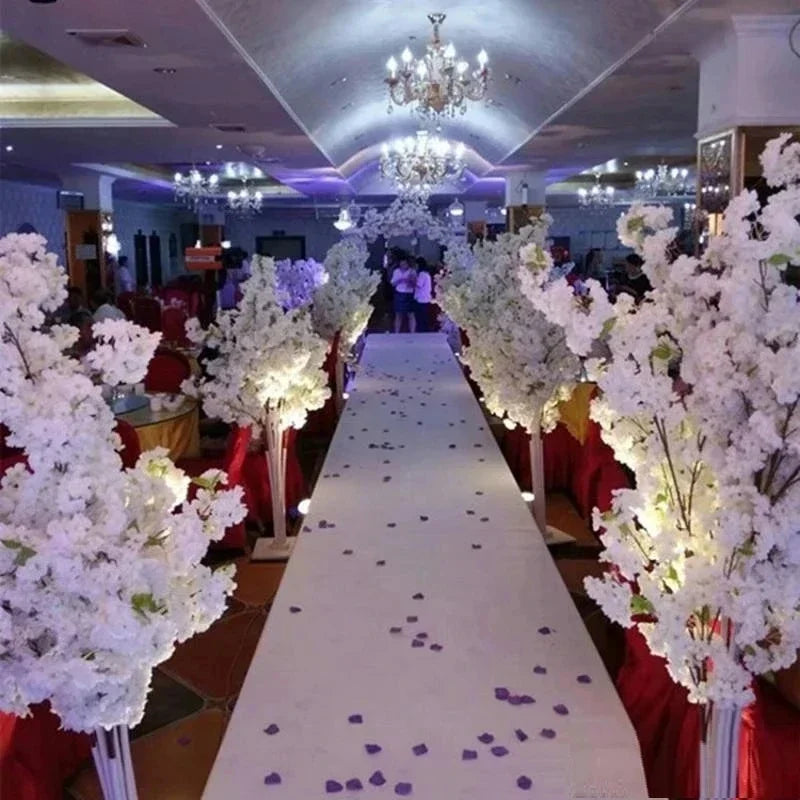 Elegant wedding aisle with white floral arrangements, illuminated by chandeliers, and scattered with purple petals on a white carpet. Red chairs line the pathway in a banquet hall.