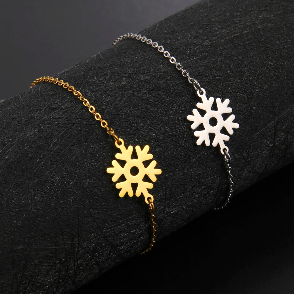 Two delicate bracelets with snowflake charms lie on a black textured surface. One bracelet is gold with a matching snowflake, while the other is silver with a silver snowflake charm.