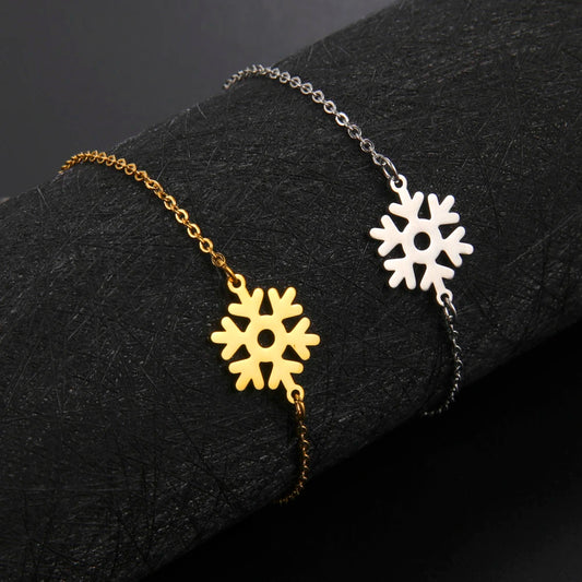 Two delicate bracelets with snowflake charms lie on a black textured surface. One bracelet is gold with a matching snowflake, while the other is silver with a silver snowflake charm.